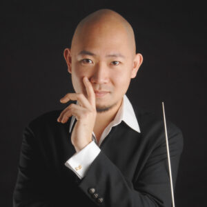Man posing in front of a dark background with one hand touching his face and a conductor's baton in the other hand