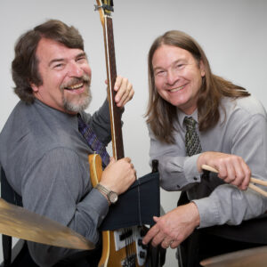 Dan and Chris Brubeck pose for a portrait. Dan holds a guitar and chris holds drum sticks.