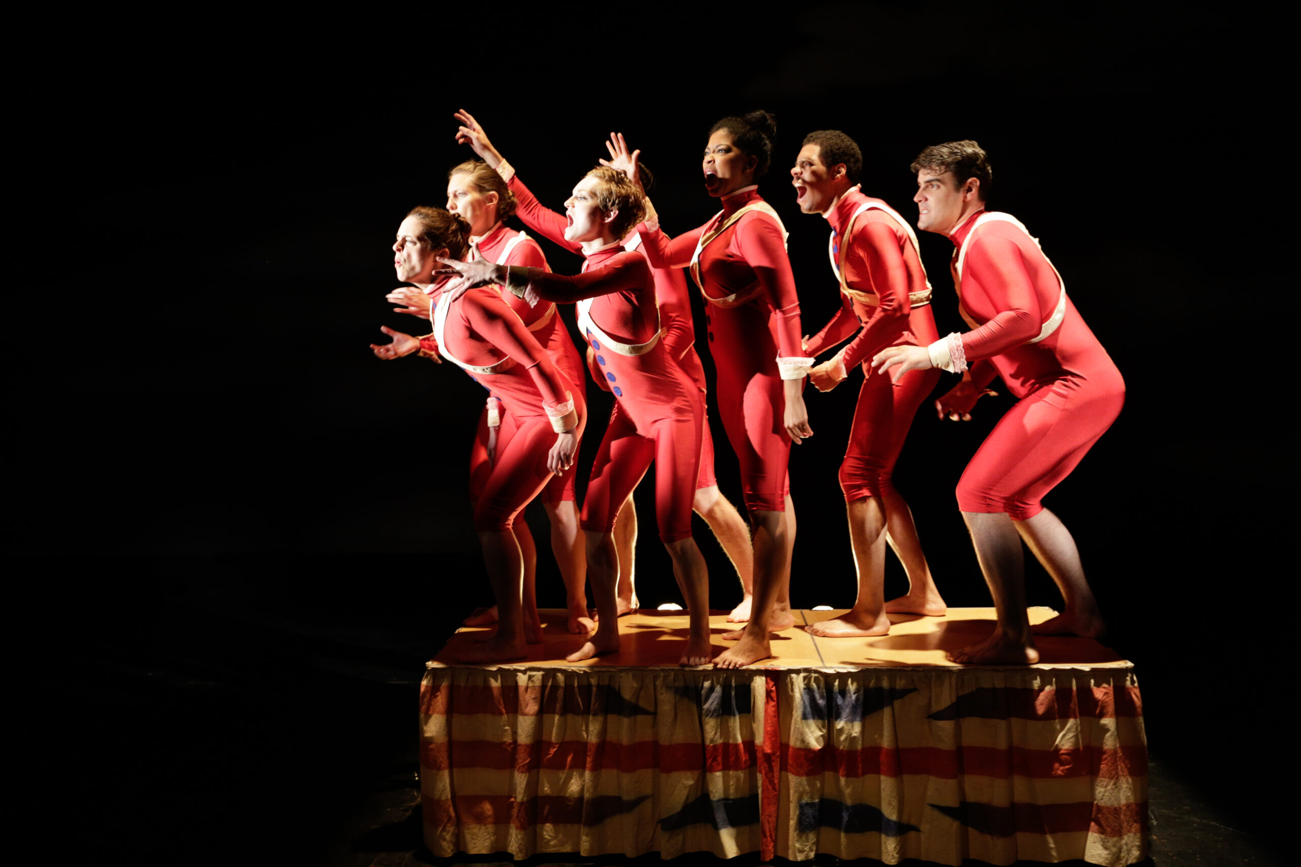 6 Actors dressed in red leotards, standing on a platform. They are all facing the left and crouching down.