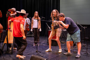 Centre students stand onstage with members of Cimarrón. One student plays the deer skull whistle and the other plays the saxophone.