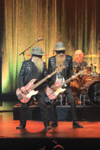 ZZ Top playing guitar on stage with their drummer in the background
