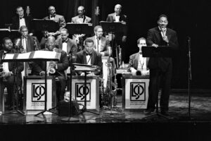 Black and white photo of Wynton Marsalis speaking into a microphone in front of the Jazz at Lincoln Center orchestra