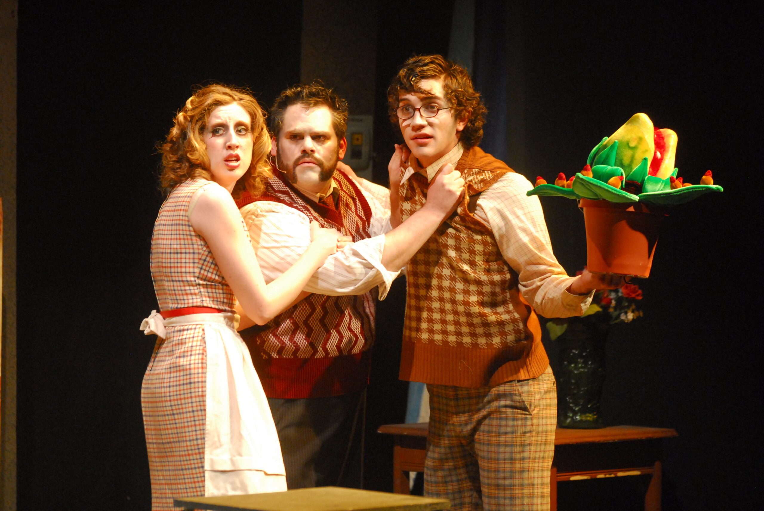 Three actors performing in Little Shop of Horrors, dressed in costume. One holds a carnivorous plant prop.