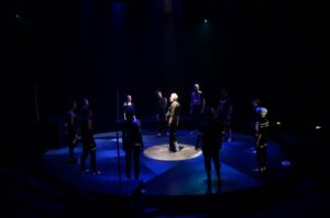 Actors stand on a dark Weisiger stage, standing in a circle around one figure in the center.