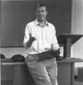 Black and white photograph of Patrick Kagan Moore standing in the front of a classroom, leaning on a desk