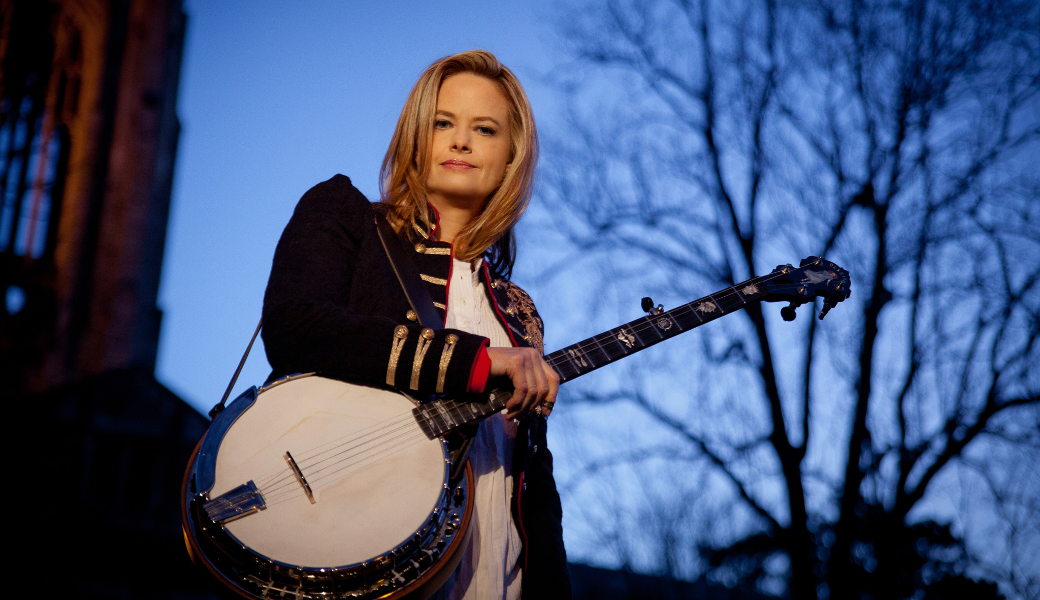 Alison Brown standing in front of a blue sky and silhouette of a tree. She is looking down at the camera and holding a banjo
