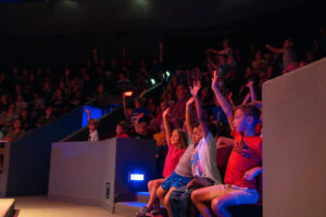 Kids sitting in a theatre, raising their hands 