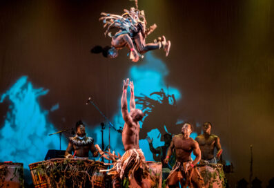 Cirque Performers On Stage. One Performer Is In The Air Spinning While Another Stands Below With His Arms Up To Catch.