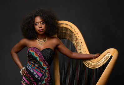 Brandee Younger Stands In Front Of Her Harp With One Arm Draped Over The Instrument