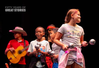 A Group Of Children Stand On Stage Holding Maracas. They Have Excited Expressions On Their Faces.