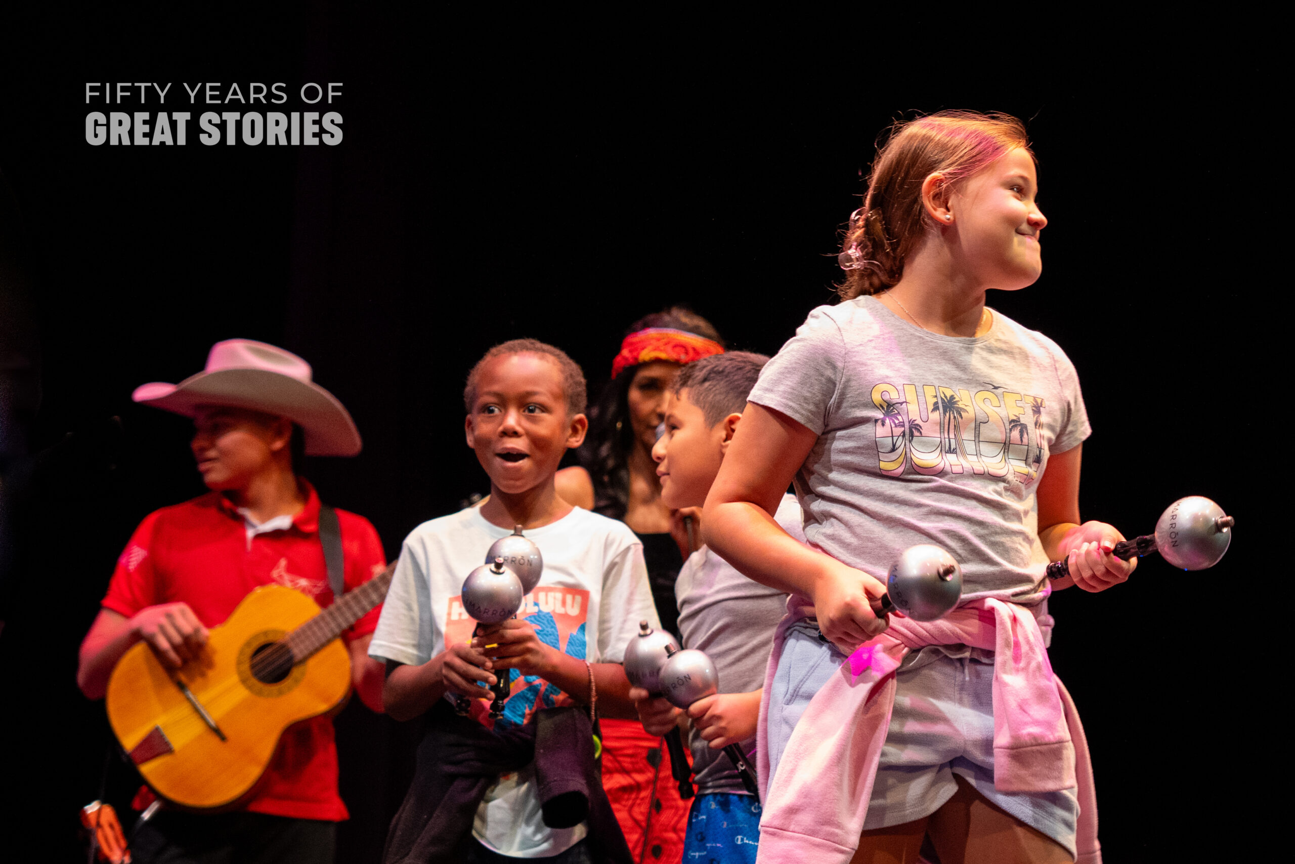 A group of children stand on stage holding maracas. They have excited expressions on their faces.