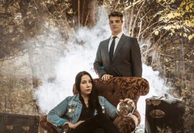 Dana And Greg Kewkirk Pose In The Woods. Smoke Is Billowing Out Behind Them. Dana Sits In A Chair And Greg Stands Behind The Chair. Both Are Staring Directly Into The Camera