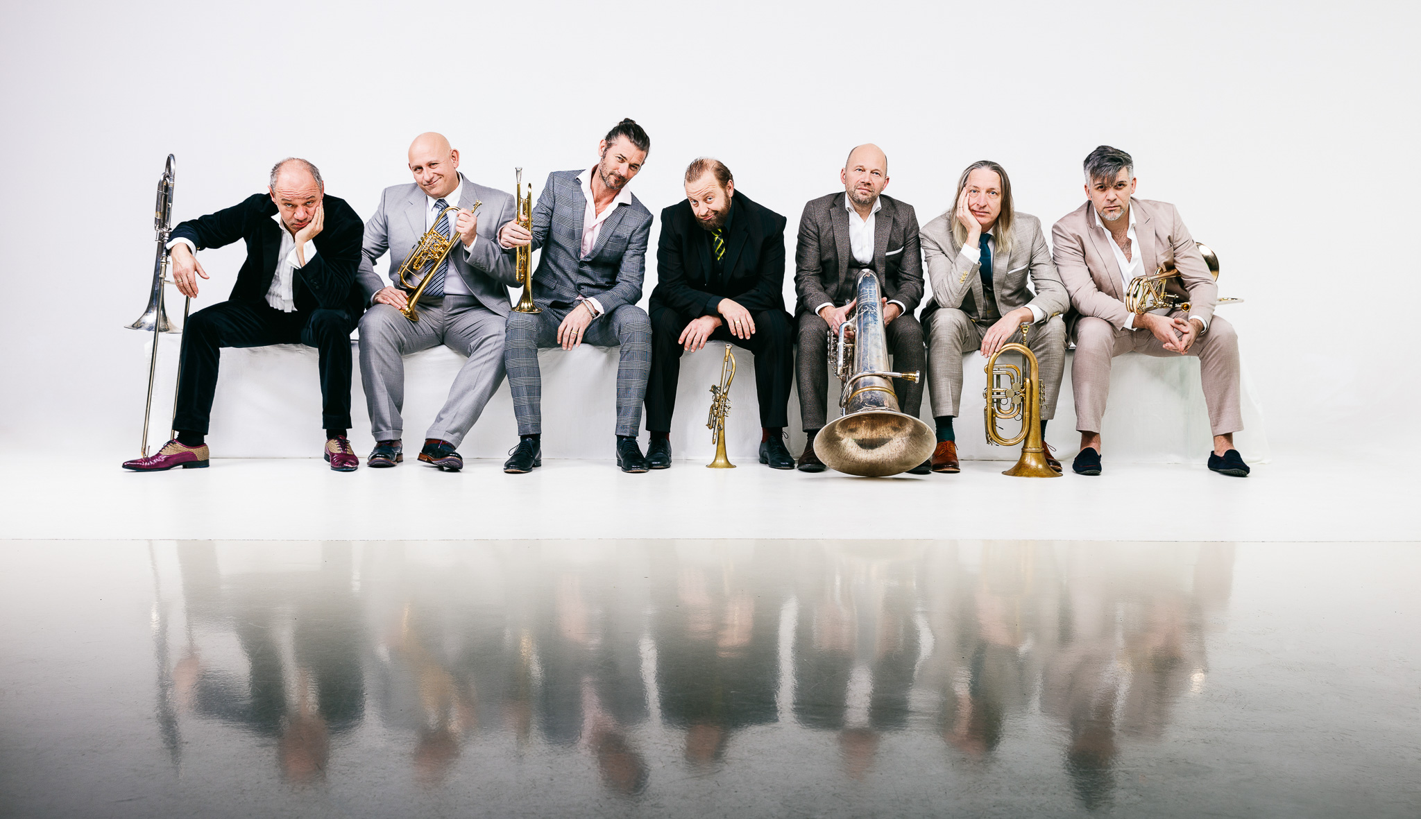 The 7 members of Mnozil Brass pose next to one another in a line in front of a white backdrop. Each holds a brass instrument.