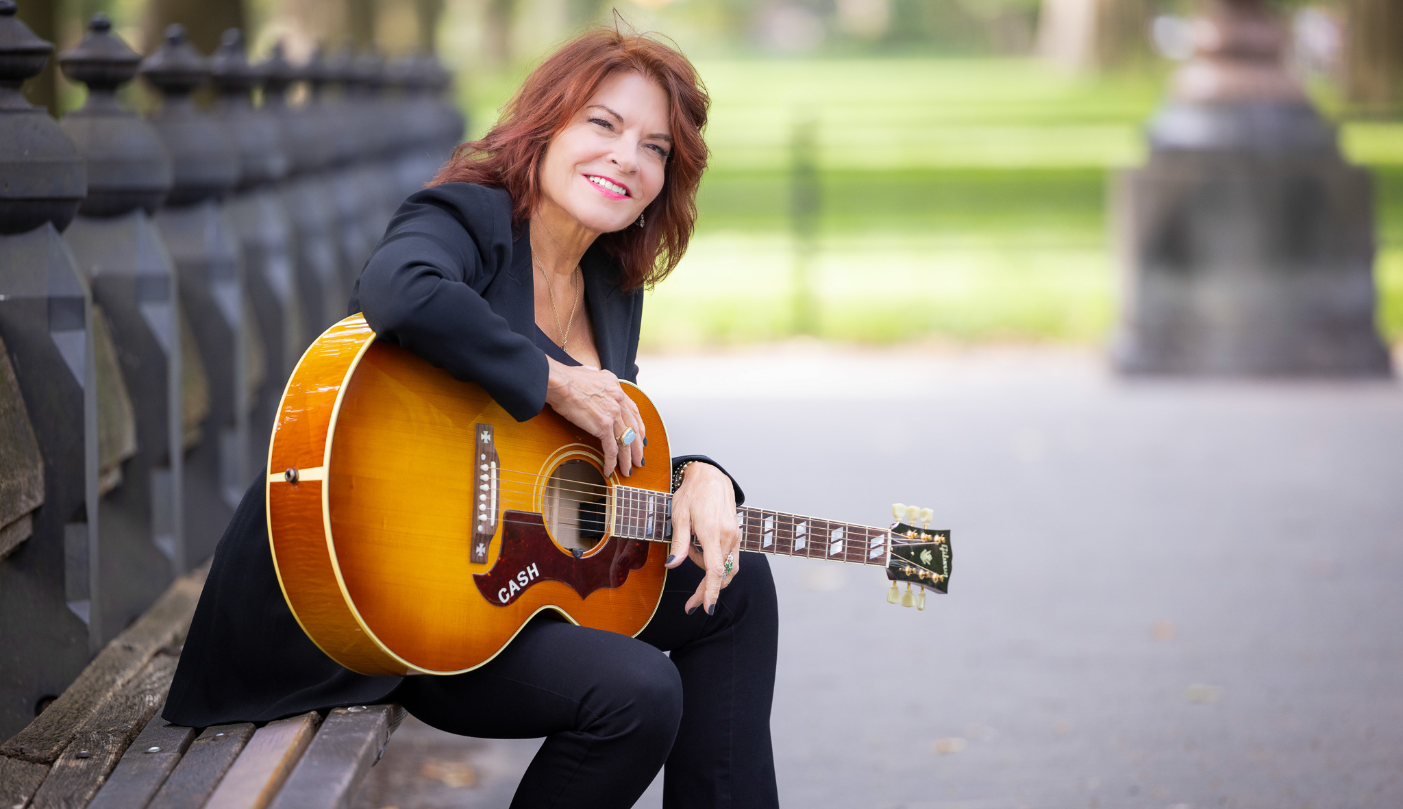 Rosanne Cash sits on a bench with a guitar on her lap and smiles toward the camera