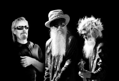 Black And White Photograph Of ZZ Top. Three Men Stand Next To Each Other. One On The Right Is Playing A Guitar