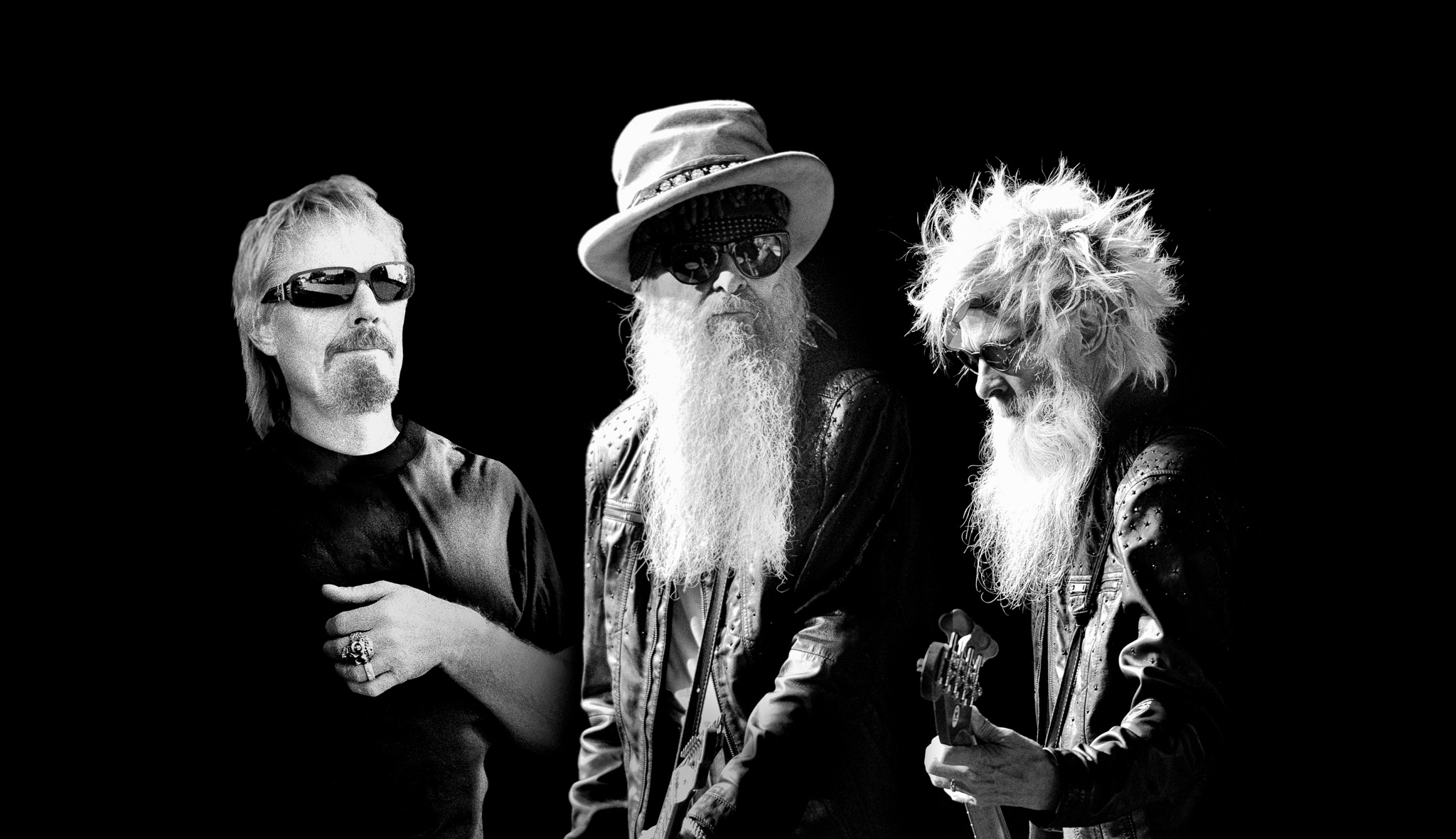 Black and white photograph of ZZ Top. Three men stand next to each other. One on the right is playing a guitar