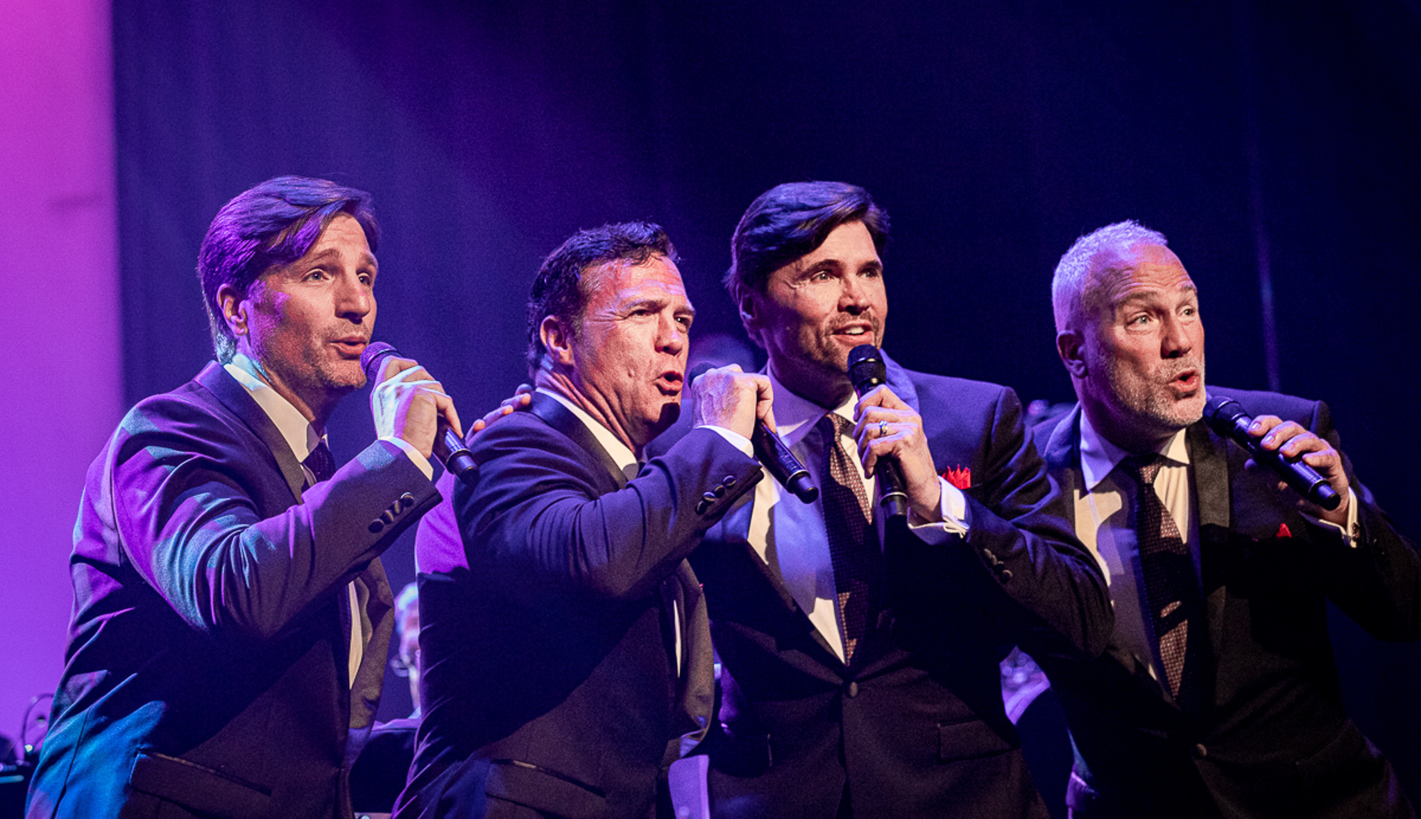 The Four Phantoms standing next to one another, performing on stage. Each is singing into a microphone
