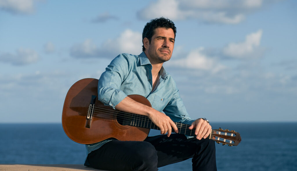Pablo Sainz Villegas sits in front of the ocean and a blue sky with his guitar resting in his lap.
