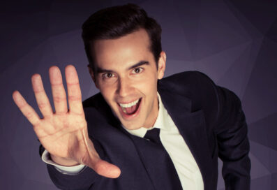 Micahel Carbonaro Wearing A Suit, Smiling Up At The Camera And Sticking His Hand Out In A Magicain-like Pose