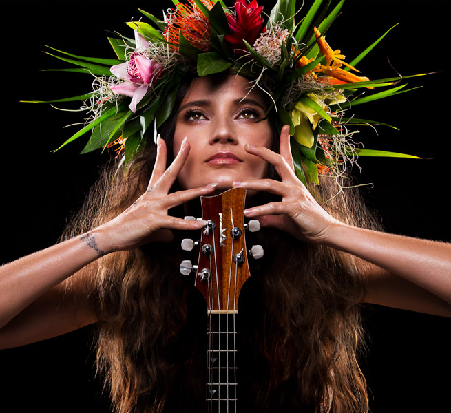 Taimane from the chest up, wearing a dramatic flower crown. Her ukulele is in front of her body and she holds her hands near her face and looks up