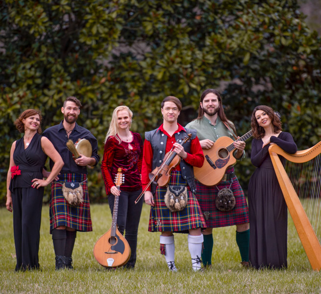 Six celtic musician band members pose in front of a tree.