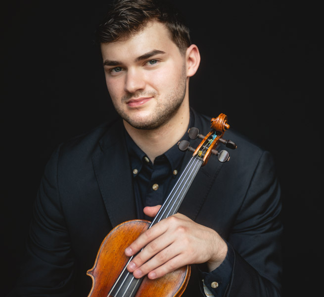 Nathan poses against a dark background. His violin lays on his lap