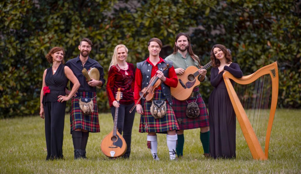 Six celtic musician band members pose in front of a tree.