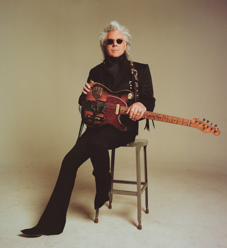 Marty Stuart, dressed in all black with sunglasses on, sits on a stool with his guitar propped in his lap
