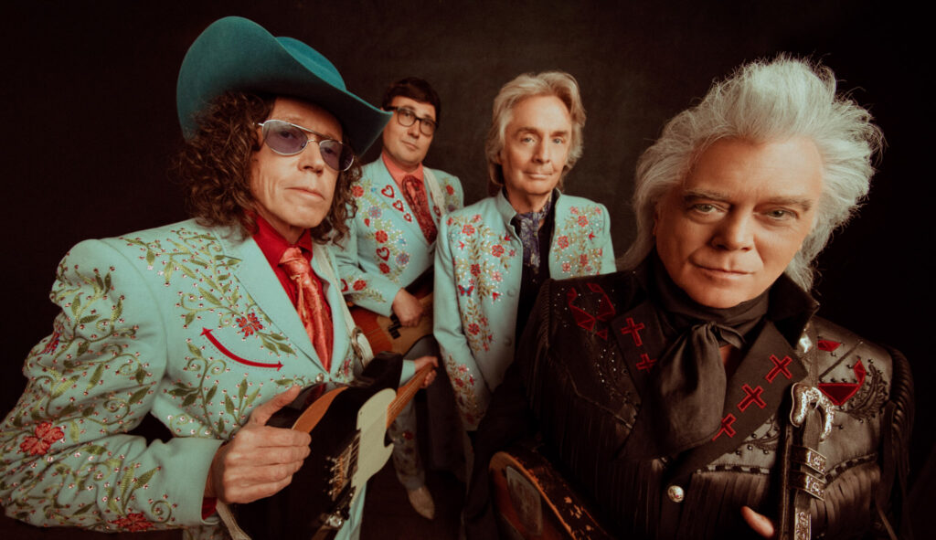 Marty Stuart and three members of his band pose against a black background. Marty wears dark colors while his band wears blue