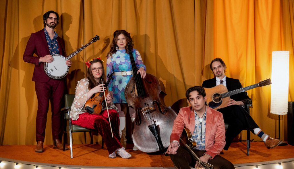 Missy Raines poses against a yellow curtain with the four other members of her band. She holds a bass propped against her hip. Her band holds a banjo, fiddle, mandolin and guitar
