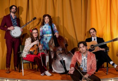 Missy Raines Poses Against A Yellow Curtain With The Four Other Members Of Her Band. She Holds A Bass Propped Against Her Hip. Her Band Holds A Banjo, Fiddle, Mandolin And Guitar