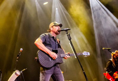 Rodney Atkins Sings Into A Microphone On Stage While Playing Guitar. Lights Shine Behind Him