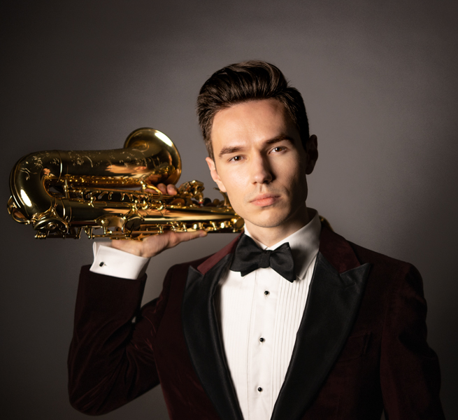 Valentin poses against a black background. He is wearing a suit and has a serious expression. He holds his saxophone up to rest on his right shoulder.