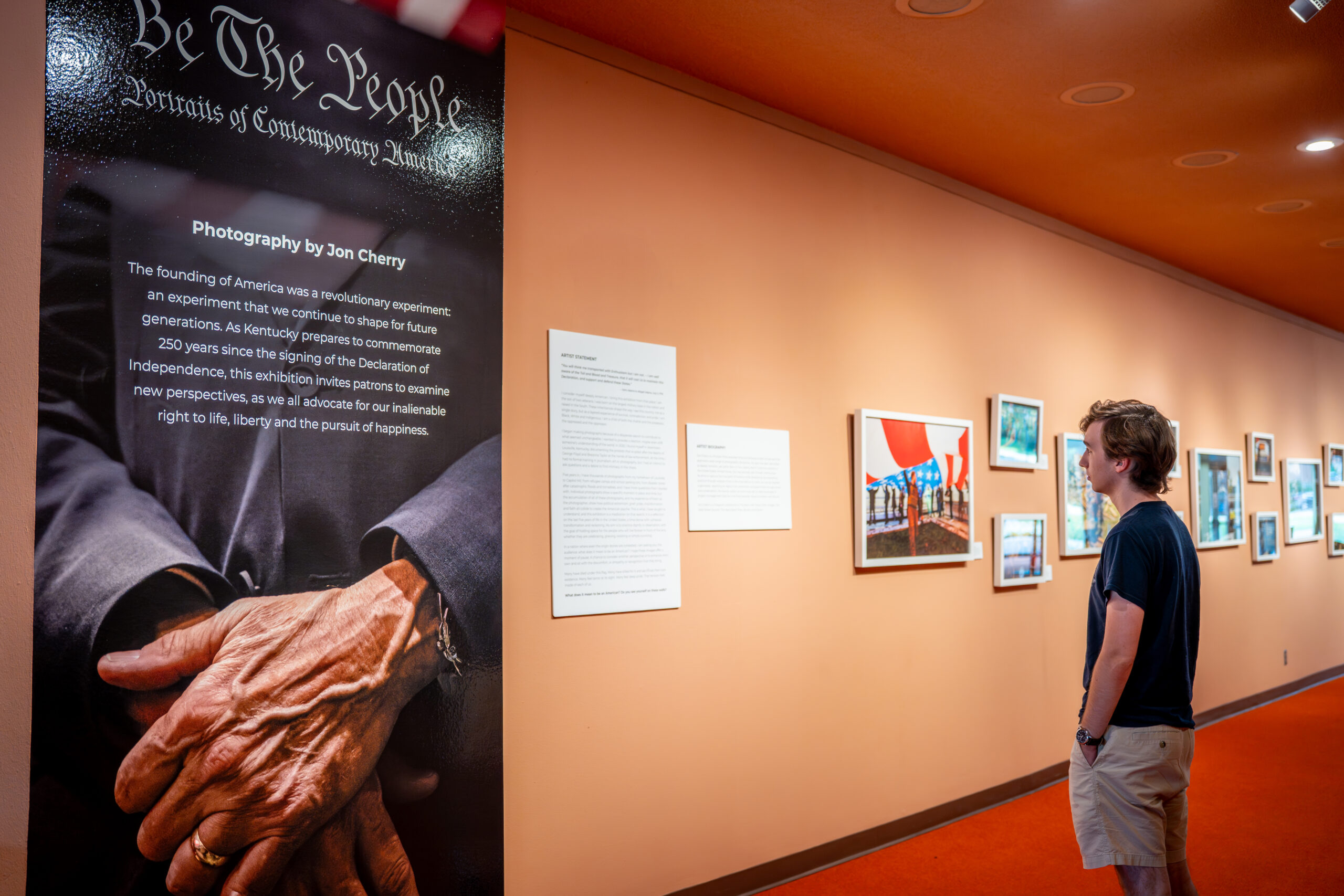 A student stands in front of a wall displaying a photography exhibition, titled "Be the People"