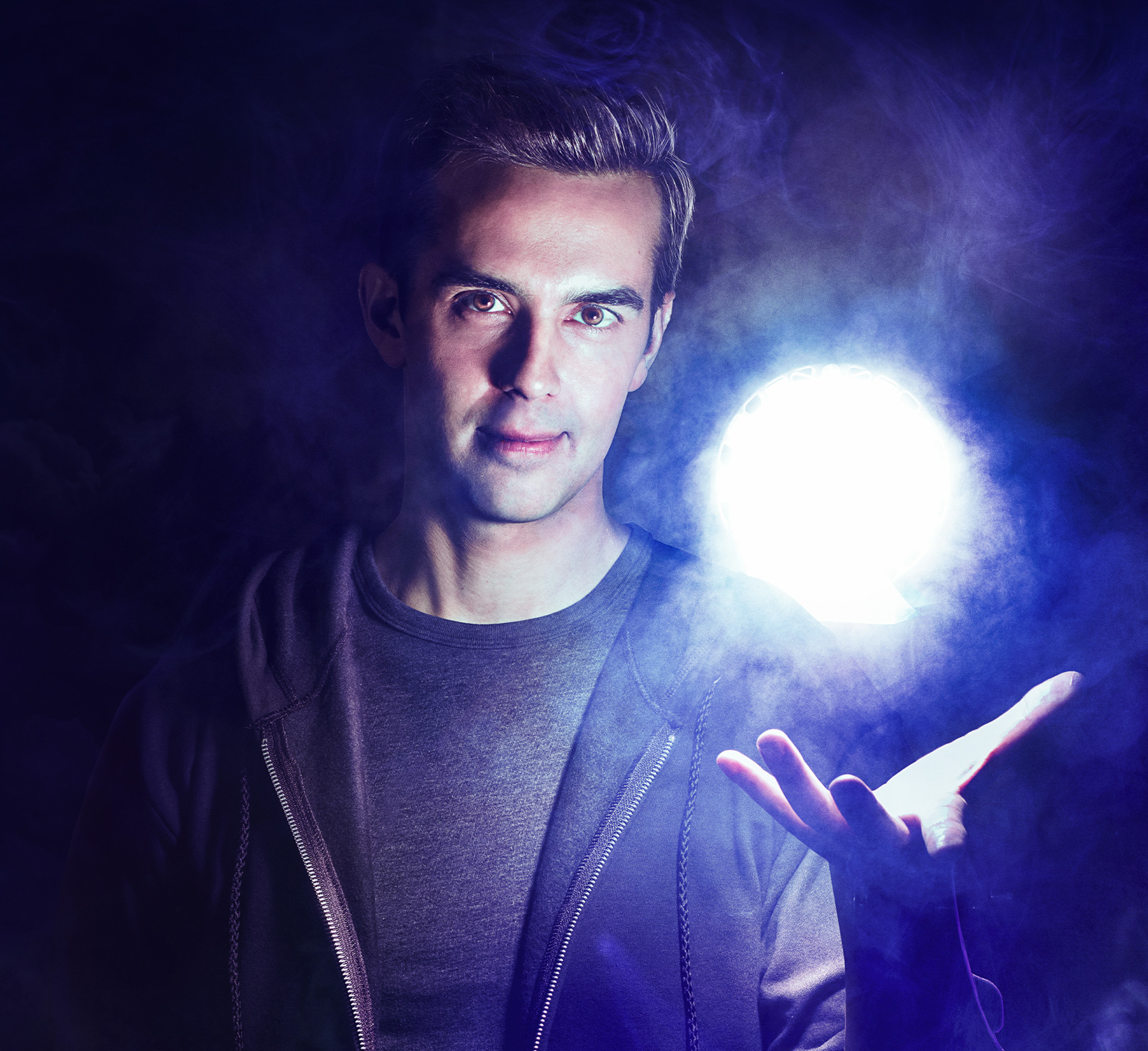 Michael Carbonaro against a dark mysterious background. He holds up his hand and a glowing orb floats above his palm