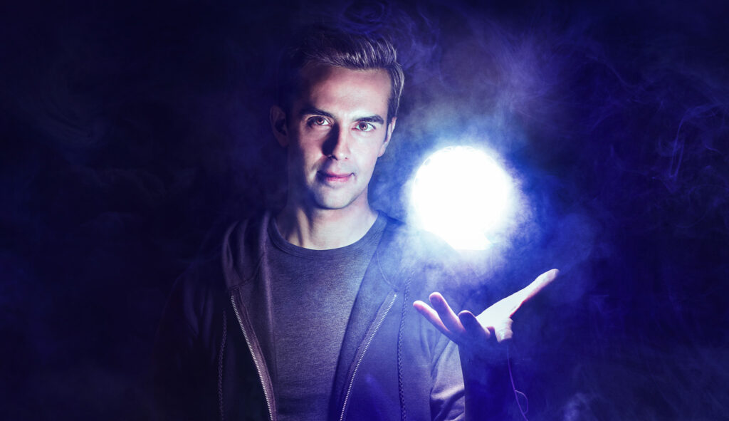 Michael Carbonaro against a dark mysterious background. He holds up his hand and a glowing orb floats above his palm