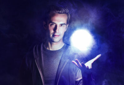Michael Carbonaro Against A Dark Mysterious Background. He Holds Up His Hand And A Glowing Orb Floats Above His Palm