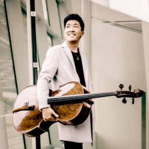 A man in a tan suit jacket holds a cello under his arm.