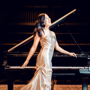 A woman in a sparkling dress stands in front of a grand piano and smiles off to the side of the frame