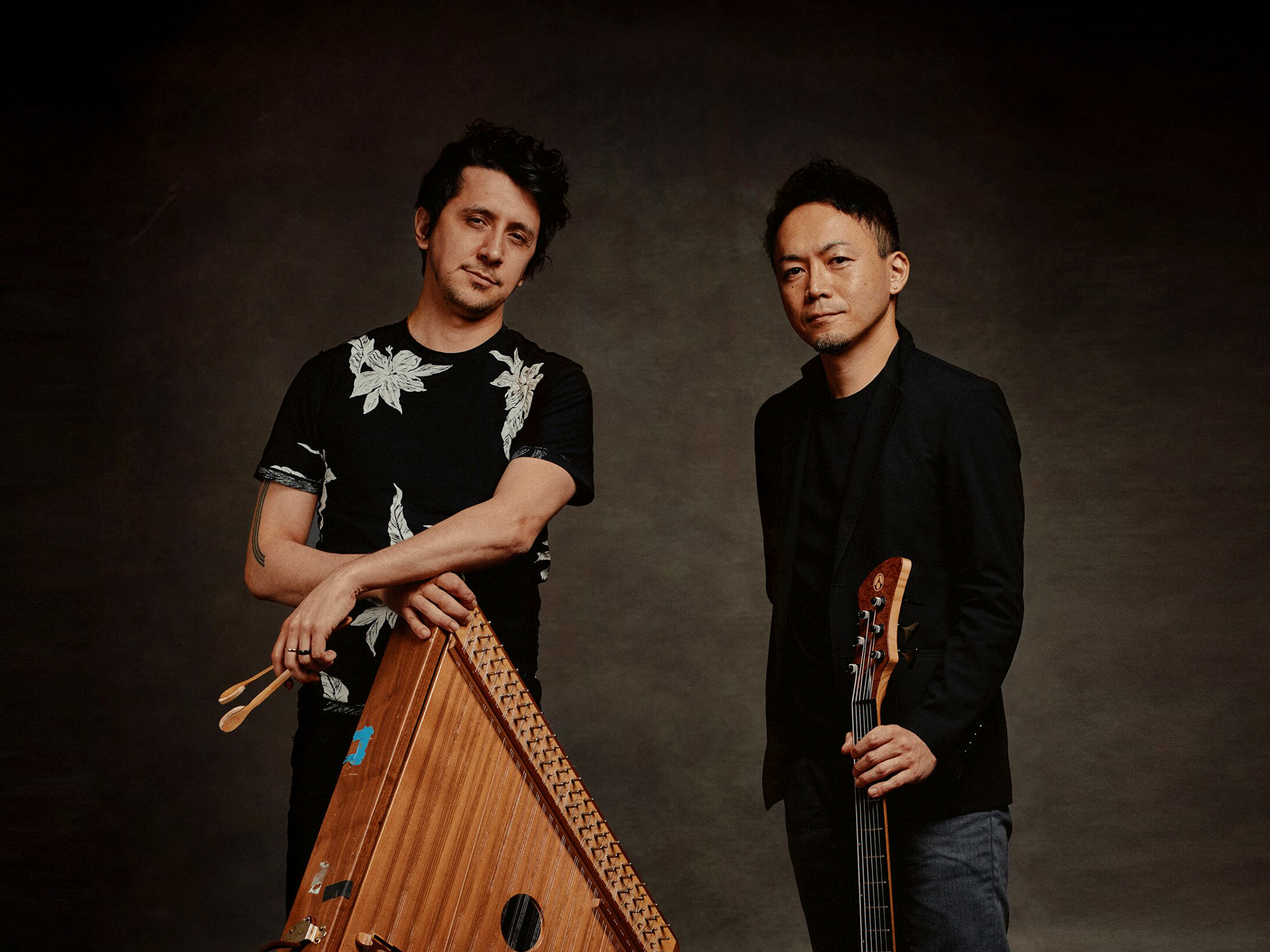 Two members of House of Waters pose against a dark background. The man on the left leans his arms against a dulcimer, and the man on the right holds a guitar