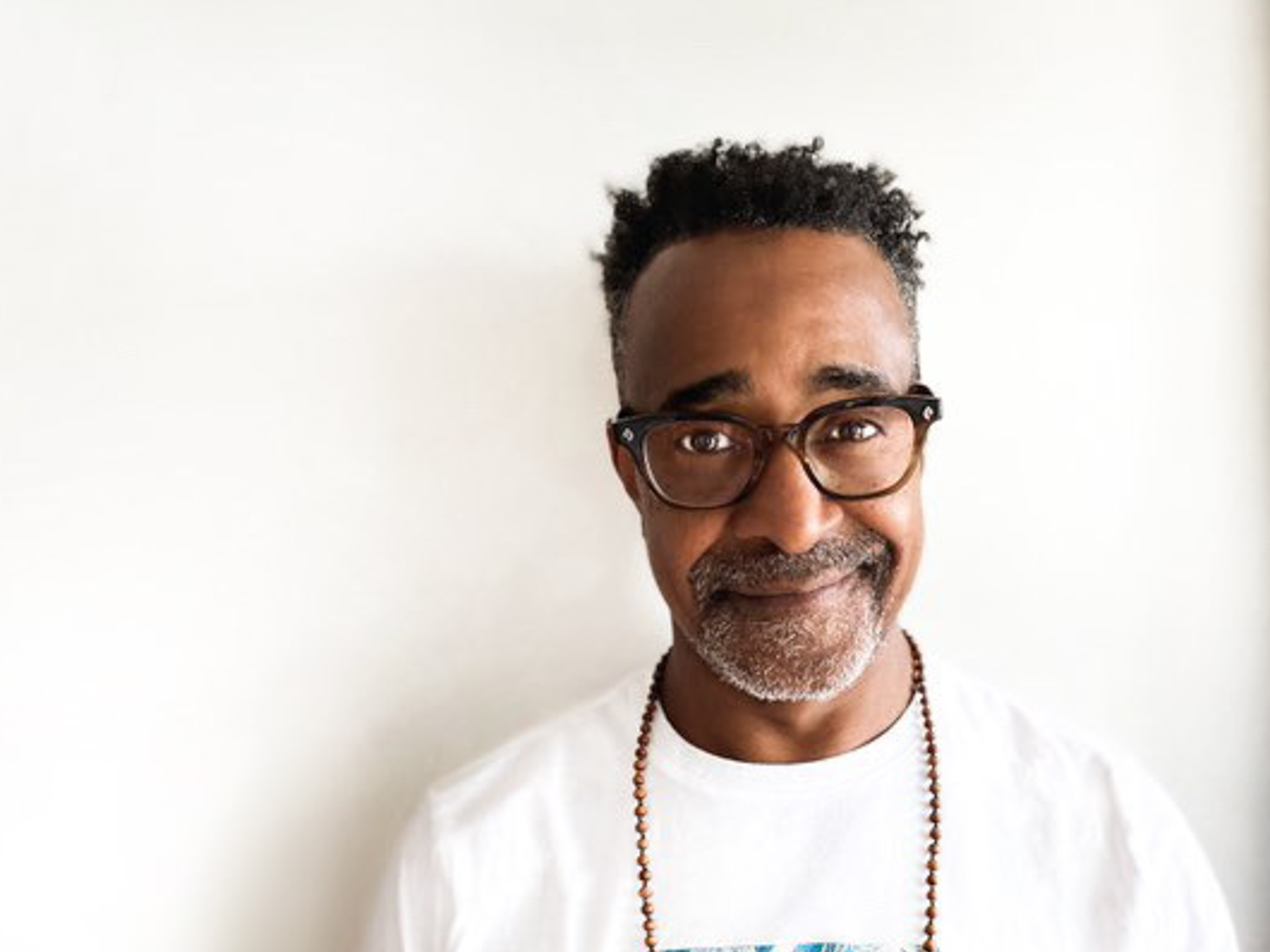 Portrait of Tim Meadows from the chest up against a white background. He is wearing glasses and a necklace and smirking at the camera