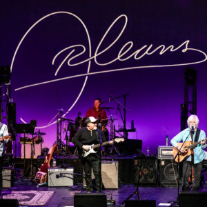 Members of Orleans play on stage with their name displayed in a large font behind them