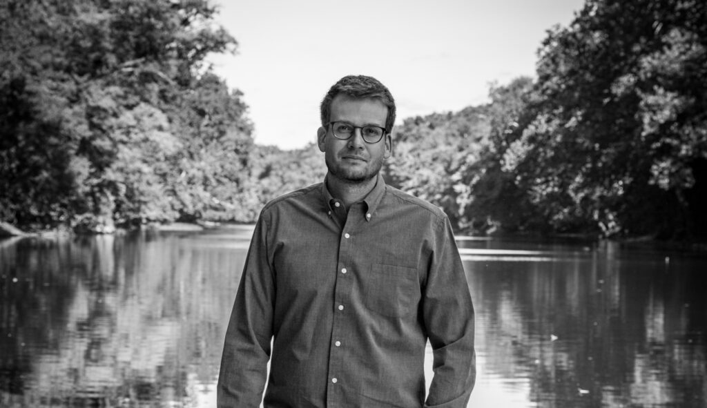 Black and white portrait of John Green standing in front of a lake