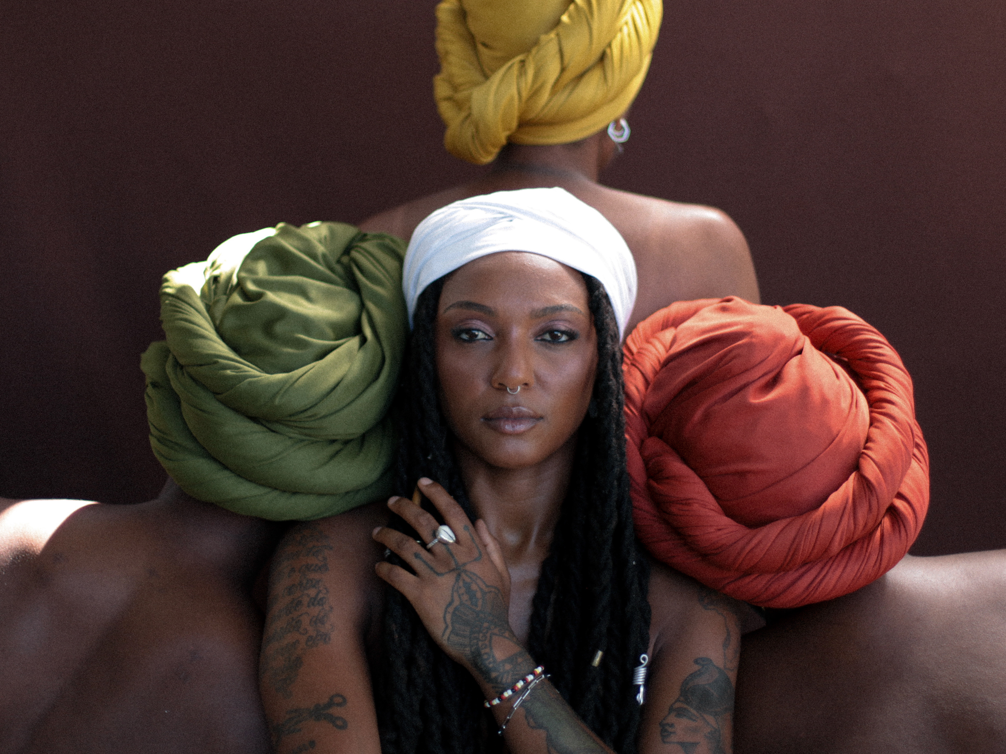 Bia Ferreira looking at the camera with a hand in front of her chest. Side by side and behind her are 3 women facing away from the camera. The women are wearing head scarves in different colors