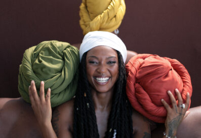 Bia Ferreira Smiling, Holding Her Hands Up. Side By Side And Behind Her Are 3 Women Facing Away From The Camera. The Women Are Wearing Head Scarves In Different Colors