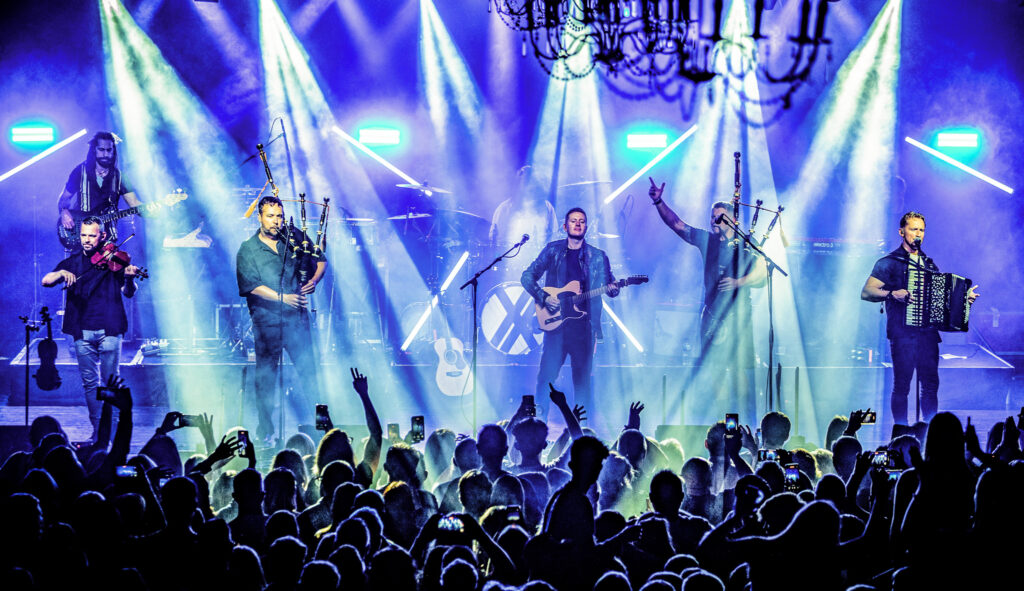 Members of the band Skerryvore performing on stage in front of a packed audience. Stage lights shine down on the performers.