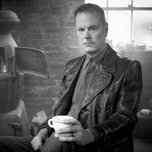 Black and white shot of Barry Bales posing by an old woodstove, holding a teacup
