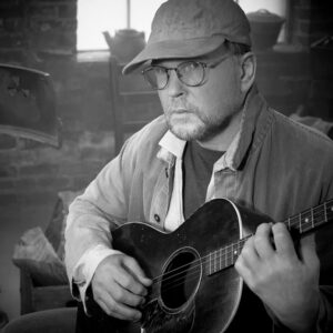 A black and white portrait of Ron Block holding a guitar