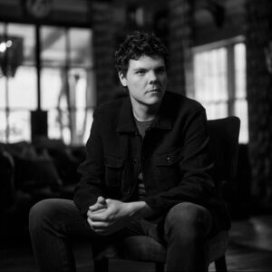 Black and white portrait of Jacob Burleson, sitting in a chair with his elbows propped on his knees