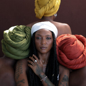 Bia Ferreira looking at the camera with a hand in front of her chest. Side by side and behind her are 3 women facing away from the camera. The women are wearing head scarves in different colors